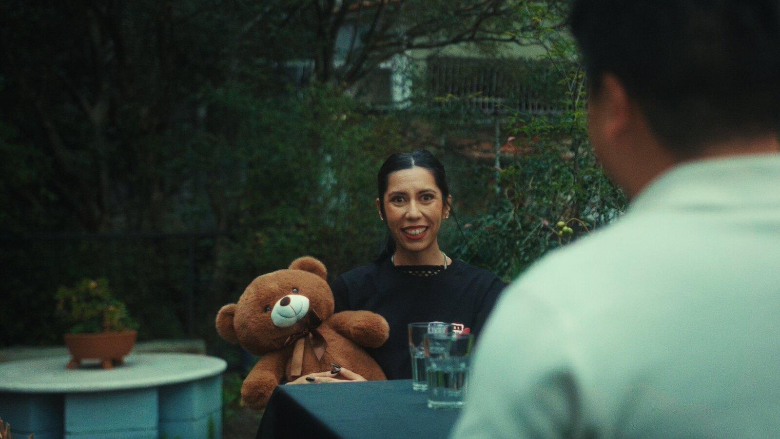 Isabella Flint sentada a uma mesa com um urso de pelúcia ao lado, em cena do clipe de Ex-amores